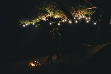 Aşık genç bir çift, ormanda bir çadırın önünde eski bir çelenkle kamp kuruyor. Çok gürültülü bir fotoğraf, seçici bir odak noktası var.