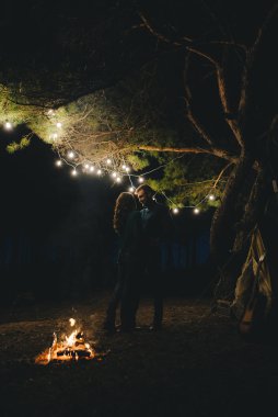 Aşık genç bir çift, ormanda bir çadırın önünde eski bir çelenkle kamp kuruyor. Çok gürültülü bir fotoğraf, seçici bir odak noktası var.