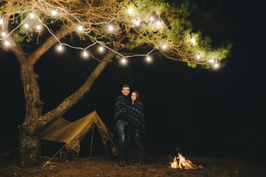 Aşık genç bir çift, ormanda bir çadırın önünde eski bir çelenkle kamp kuruyor. Çok gürültülü bir fotoğraf, seçici bir odak noktası var.