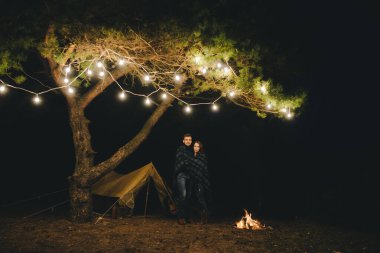 Aşık genç bir çift, ormanda bir çadırın önünde eski bir çelenkle kamp kuruyor. Çok gürültülü bir fotoğraf, seçici bir odak noktası var.