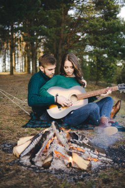 Aşık genç bir çift kamp ateşi başında çadır kurmuş gitar çalıyor.