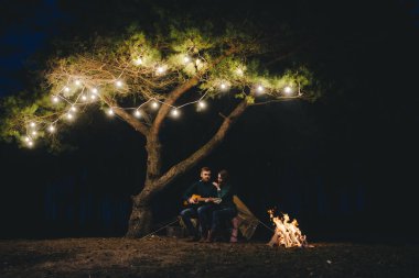 Aşık genç bir çift, ormanda bir çadırın önünde eski bir çelenkle kamp kuruyor. Çok gürültülü bir fotoğraf, seçici bir odak noktası var.