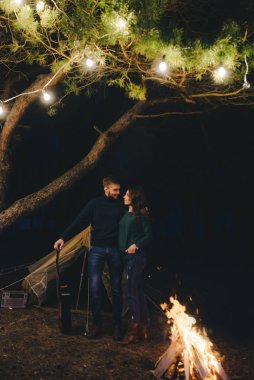 Aşık genç bir çift, ormanda bir çadırın önünde eski bir çelenkle kamp kuruyor. Çok gürültülü bir fotoğraf, seçici bir odak noktası var.