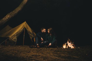 Aşık genç bir çift, ormanda bir çadırın önünde eski bir çelenkle kamp kuruyor. Çok gürültülü bir fotoğraf, seçici bir odak noktası var.