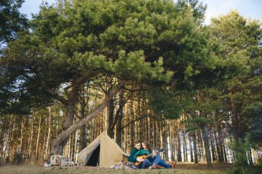Aşık genç bir çift kamp ateşi başında çadır kurmuş gitar çalıyor.