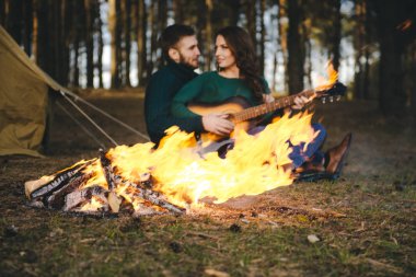 Aşık genç bir çift kamp ateşi başında çadır kurmuş gitar çalıyor.