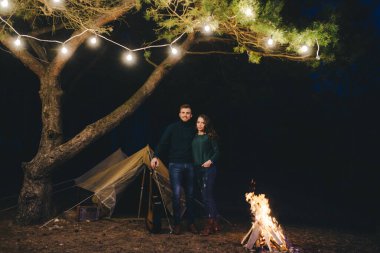 Aşık genç bir çift, ormanda bir çadırın önünde eski bir çelenkle kamp kuruyor. Çok gürültülü bir fotoğraf, seçici bir odak noktası var.