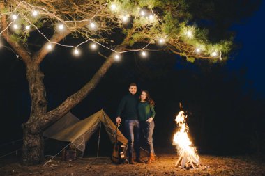 Aşık genç bir çift, ormanda bir çadırın önünde eski bir çelenkle kamp kuruyor. Çok gürültülü bir fotoğraf, seçici bir odak noktası var.