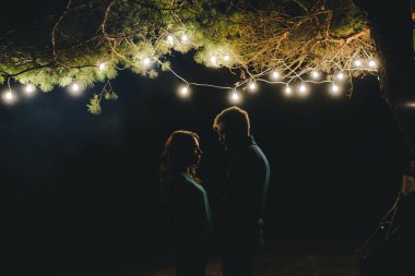 Aşık genç bir çift, ormanda bir çadırın önünde eski bir çelenkle kamp kuruyor. Çok gürültülü bir fotoğraf, seçici bir odak noktası var.