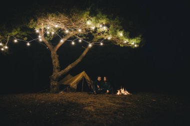 Aşık genç bir çift, ormanda bir çadırın önünde eski bir çelenkle kamp kuruyor. Çok gürültülü bir fotoğraf, seçici bir odak noktası var.