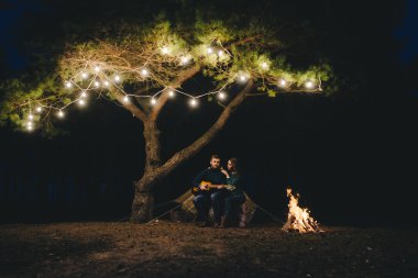 Aşık genç bir çift, ormanda bir çadırın önünde eski bir çelenkle kamp kuruyor. Çok gürültülü bir fotoğraf, seçici bir odak noktası var.