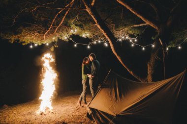 Aşık genç bir çift, ormanda bir çadırın önünde eski bir çelenkle kamp kuruyor. Çok gürültülü bir fotoğraf, seçici bir odak noktası var.