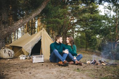 Aşık genç bir çift, kamp yapan turistleri ormandaki bir çadırın önünde oturup retro radyo dinliyorlar.