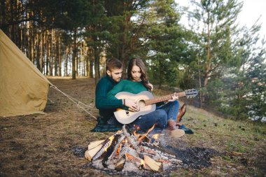 Aşık genç bir çift kamp ateşi başında çadır kurmuş gitar çalıyor.