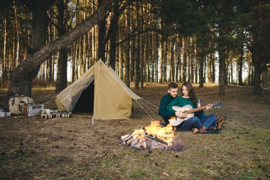 Aşık genç bir çift kamp ateşi başında çadır kurmuş gitar çalıyor.