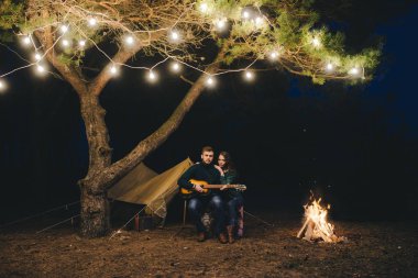 Aşık genç bir çift, ormanda bir çadırın önünde eski bir çelenkle kamp kuruyor. Çok gürültülü bir fotoğraf, seçici bir odak noktası var.