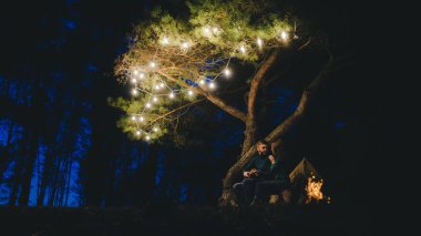 Aşık genç bir çift, ormanda bir çadırın önünde eski bir çelenkle kamp kuruyor. Çok gürültülü bir fotoğraf, seçici bir odak noktası var.
