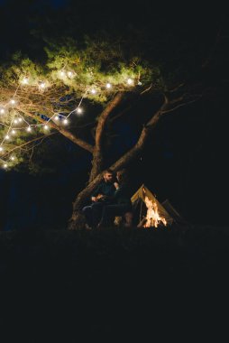 Aşık genç bir çift, ormanda bir çadırın önünde eski bir çelenkle kamp kuruyor. Çok gürültülü bir fotoğraf, seçici bir odak noktası var.