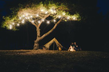 Aşık genç bir çift, ormanda bir çadırın önünde eski bir çelenkle kamp kuruyor. Çok gürültülü bir fotoğraf, seçici bir odak noktası var.