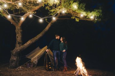 Aşık genç bir çift, ormanda bir çadırın önünde eski bir çelenkle kamp kuruyor. Çok gürültülü bir fotoğraf, seçici bir odak noktası var.