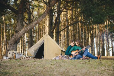 Aşık genç bir çift kamp ateşi başında çadır kurmuş gitar çalıyor.