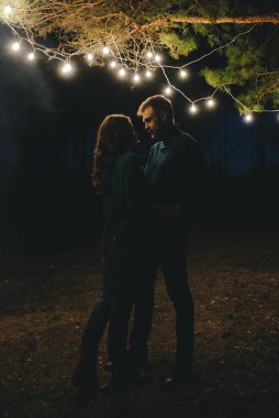 Aşık genç bir çift, ormanda bir çadırın önünde eski bir çelenkle kamp kuruyor. Çok gürültülü bir fotoğraf, seçici bir odak noktası var.