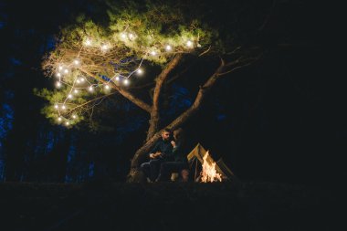 Aşık genç bir çift, ormanda bir çadırın önünde eski bir çelenkle kamp kuruyor. Çok gürültülü bir fotoğraf, seçici bir odak noktası var.