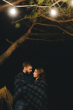 Aşık genç bir çift, ormanda bir çadırın önünde eski bir çelenkle kamp kuruyor. Çok gürültülü bir fotoğraf, seçici bir odak noktası var.