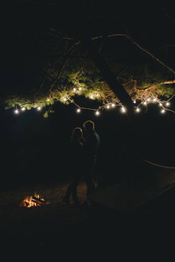 Aşık genç bir çift, ormanda bir çadırın önünde eski bir çelenkle kamp kuruyor. Çok gürültülü bir fotoğraf, seçici bir odak noktası var.