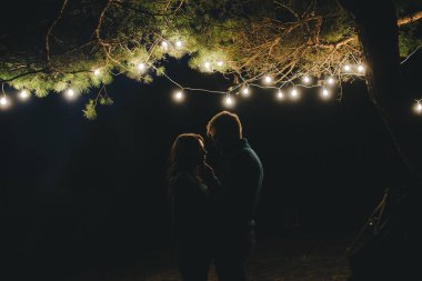 Aşık genç bir çift, ormanda bir çadırın önünde eski bir çelenkle kamp kuruyor. Çok gürültülü bir fotoğraf, seçici bir odak noktası var.