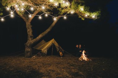 Aşık genç bir çift, ormanda bir çadırın önünde eski bir çelenkle kamp kuruyor. Çok gürültülü bir fotoğraf, seçici bir odak noktası var.