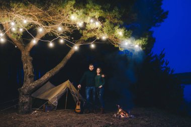 Aşık genç bir çift, ormanda bir çadırın önünde eski bir çelenkle kamp kuruyor. Çok gürültülü bir fotoğraf, seçici bir odak noktası var.