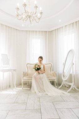 Parlak bir odada romantik bir ortamda genç ve güzel bir gelinin portresi. Gecelikli gelin ve düğün buketi.