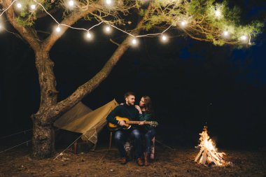 Aşık genç bir çift, ormanda bir çadırın önünde eski bir çelenkle kamp kuruyor. Çok gürültülü bir fotoğraf, seçici bir odak noktası var.