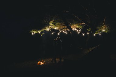 Aşık genç bir çift, ormanda bir çadırın önünde eski bir çelenkle kamp kuruyor. Çok gürültülü bir fotoğraf, seçici bir odak noktası var.