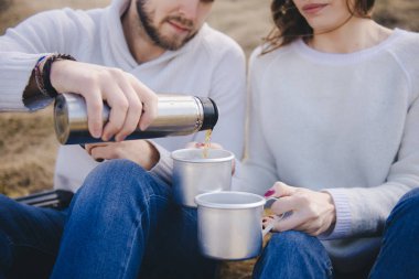 Turist çantalı ve gitarlı mutlu kız ve adam bir tarlada oturup termostan çay içiyorlar, seyahat aşkı konsepti.