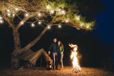 Aşık genç bir çift, ormanda bir çadırın önünde eski bir çelenkle kamp kuruyor. Çok gürültülü bir fotoğraf, seçici bir odak noktası var.