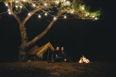 Aşık genç bir çift, ormanda bir çadırın önünde eski bir çelenkle kamp kuruyor. Çok gürültülü bir fotoğraf, seçici bir odak noktası var.