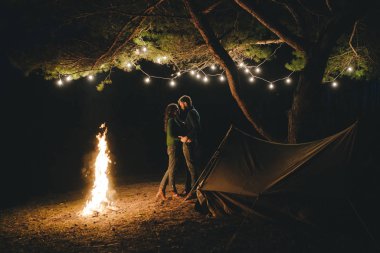 Aşık genç bir çift, ormanda bir çadırın önünde eski bir çelenkle kamp kuruyor. Çok gürültülü bir fotoğraf, seçici bir odak noktası var.