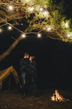 Aşık genç bir çift, ormanda bir çadırın önünde eski bir çelenkle kamp kuruyor. Çok gürültülü bir fotoğraf, seçici bir odak noktası var.