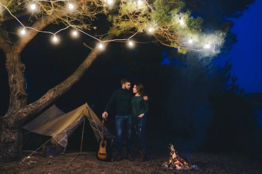 Aşık genç bir çift, ormanda bir çadırın önünde eski bir çelenkle kamp kuruyor. Çok gürültülü bir fotoğraf, seçici bir odak noktası var.