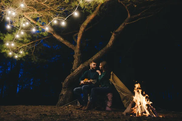 Young couple in love camping tourists sitting by a fire against a tent in the forest with a retro garland, photo with a lot of noise, selective focus