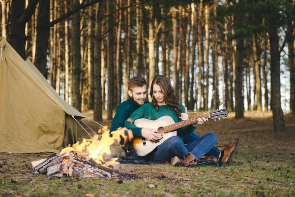 Aşık genç bir çift kamp ateşi başında çadır kurmuş gitar çalıyor.