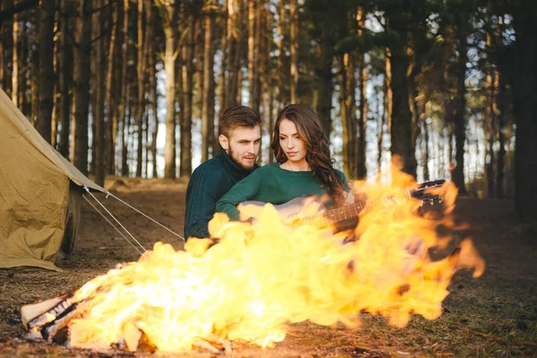 Aşık genç bir çift kamp ateşi başında çadır kurmuş gitar çalıyor.