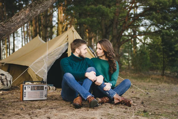 森の中のテントに対して火のそばに座ってレトロなラジオを聞く愛のキャンプ観光客の若いカップル — ストック写真