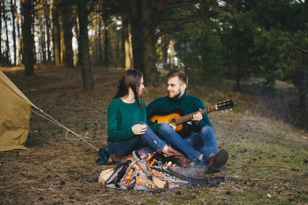 Aşık genç bir çift kamp ateşi başında çadır kurmuş gitar çalıyor.