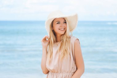 Şapkalı ve uzun elbiseli çekici sarışın bir kadın masmavi deniz kıyısındaki kar beyazı kumların üzerinde duruyor.