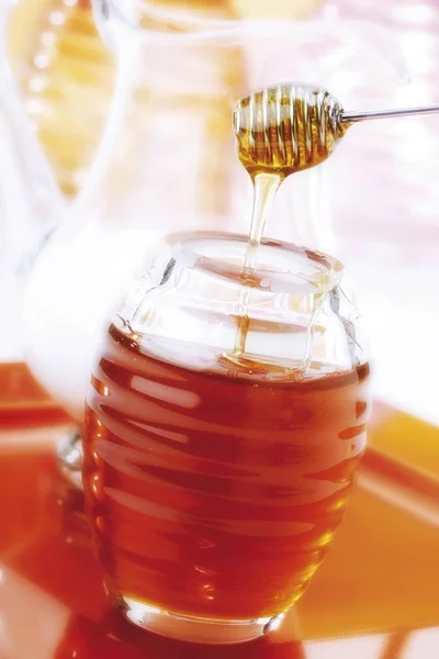 Honey dripping from honey dripper into a jar of honey, jug of milk in the background