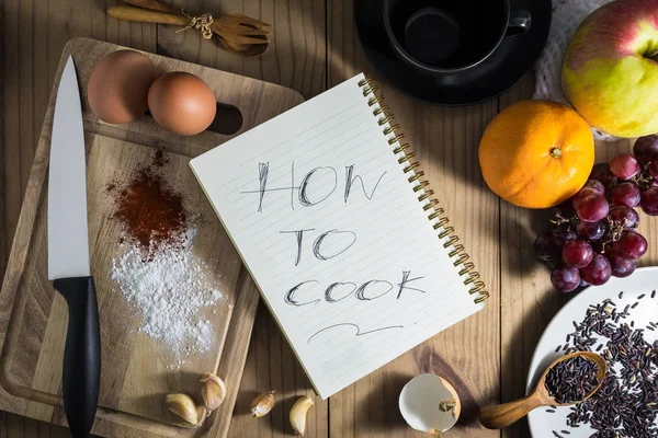 Вид зверху на приготування їжі: білий блокнот має літеру "HOW TO COOK " і яйця, ніж, ароматизатор на рубаній дошці. і чашка чаю, фрукти, ложка, виделка на дерев'яному столі. і коричневий рис на білій тарілці . — стокове фото