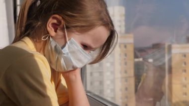 Karantina, koronavirüs tehdidi. Koruyucu tıbbi maskeli üzgün çocuk pencere eşiğinde oturur ve pencereden dışarı bakar. Virüs koruması, salgın hastalık, önleme salgını.